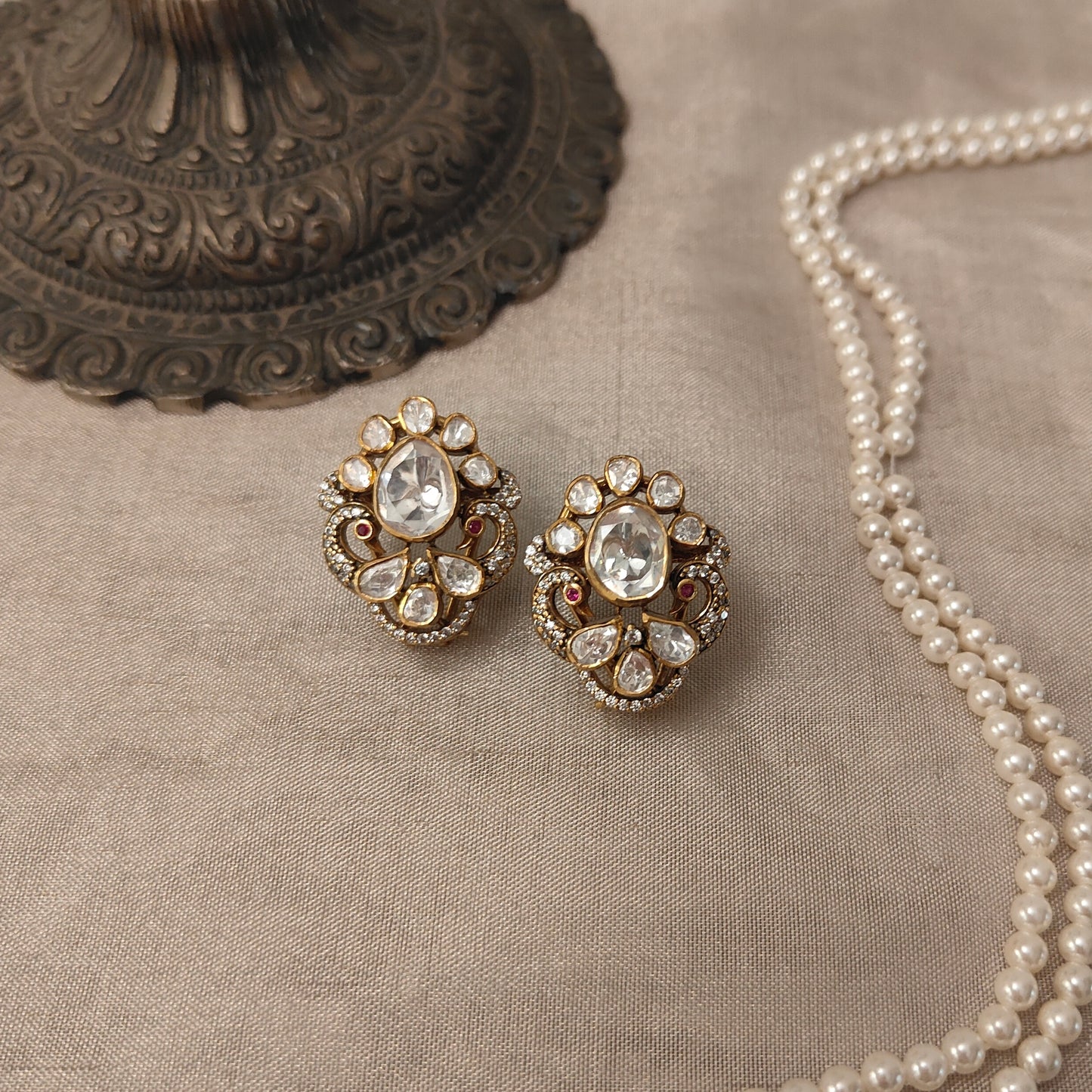 "Gold-plated polki Noor Classic stud earrings with oval centerpiece and floral crystal design by Arka Jaipur", "Traditional kundan-style stud earrings with polki stones and ruby accents","Designer Indian stud earrings with floral motif and diamond-style detailing","Luxury heritage stud earrings handcrafted in Jaipur with polki and crystal work"