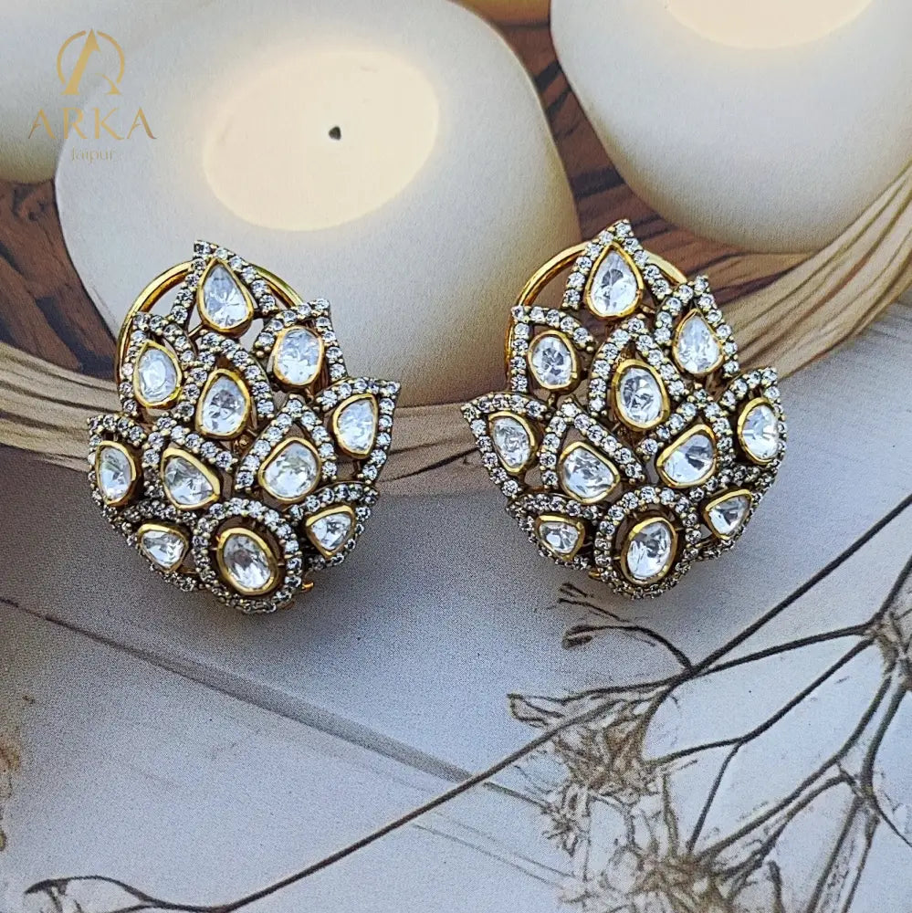 These exquisite Kamalika earrings showcase a stunning floral-inspired cluster design, crafted with teardrop-shaped stones arranged in a layered petal motif. Set in a rich gold-tone Sterling Silver base and bordered with delicate pavé crystals, the design radiates elegance and grandeur. Perfect for weddings, festive occasions, and evening celebrations, these statement studs add timeless charm and brilliance to any look.