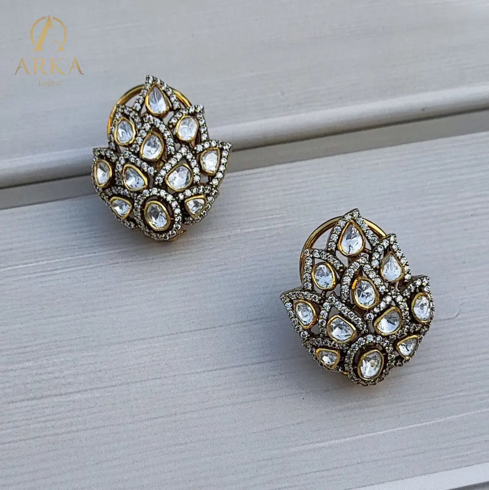 These exquisite Kamalika earrings showcase a stunning floral-inspired cluster design, crafted with teardrop-shaped stones arranged in a layered petal motif. Set in a rich gold-tone Sterling Silver base and bordered with delicate pavé crystals, the design radiates elegance and grandeur. Perfect for weddings, festive occasions, and evening celebrations, these statement studs add timeless charm and brilliance to any look.