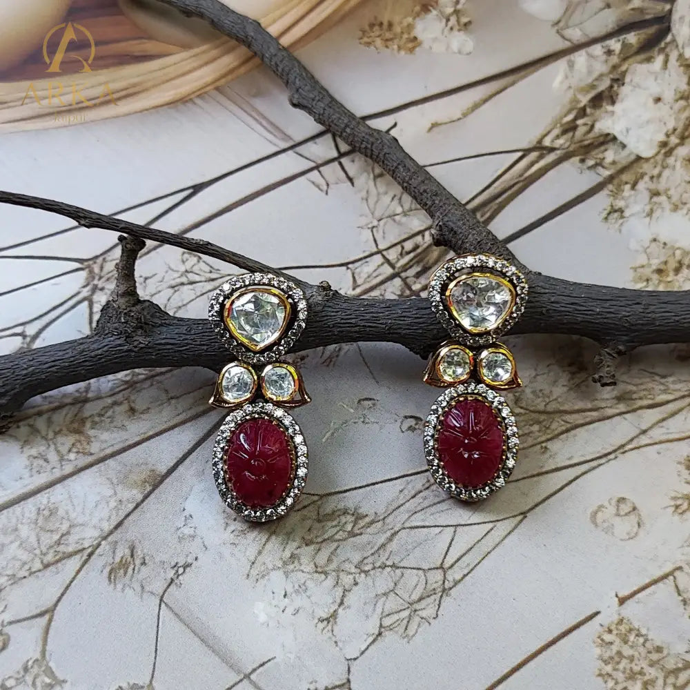 Regal Ruby earrings by Arka Jaipur, handcrafted gold-plated silver earrings with carved ruby red centerpieces, moissanite polki accents, and CZ diamond halo, displayed on a rustic branch with floral background.