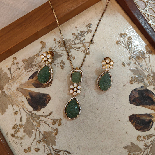 "Elysian Emerald Drop Pendant Set Handcrafted  by Arka Jaipur with carved green gemstone top and teardrop Polki stone drop, outlined with CZ stones, displayed on a wooden background"