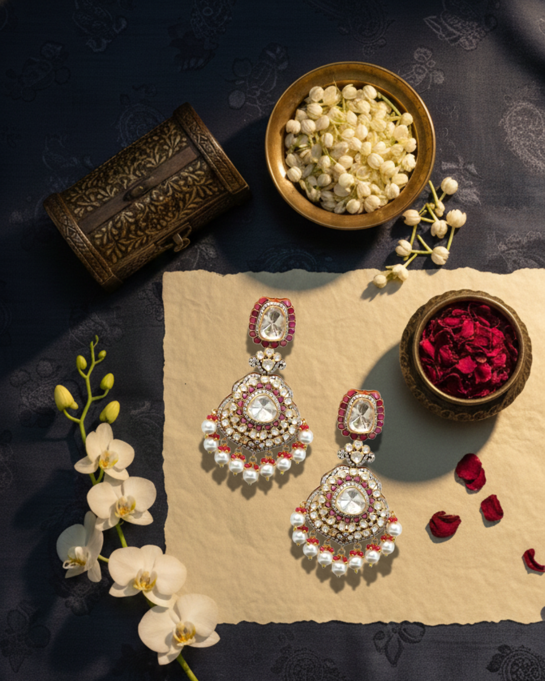 The Rosetta Royale earrings with pearls and red stones on a dark fabric background with flowers and a box.