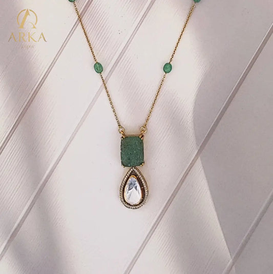 "Elysian Emerald Drops Pendant Handcrafted  by Arka Jaipur with carved green gemstone top and teardrop Polki stone drop, outlined with CZ stones, displayed on a wooden background"