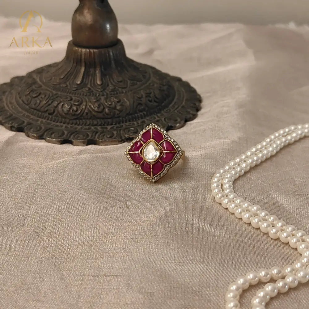 “Lotus Bloom Ring by Arka Jaipur featuring ruby enamel lotus petals and a central polki-style stone, placed on a beige fabric with a vintage brass decor piece and pearl necklace.”