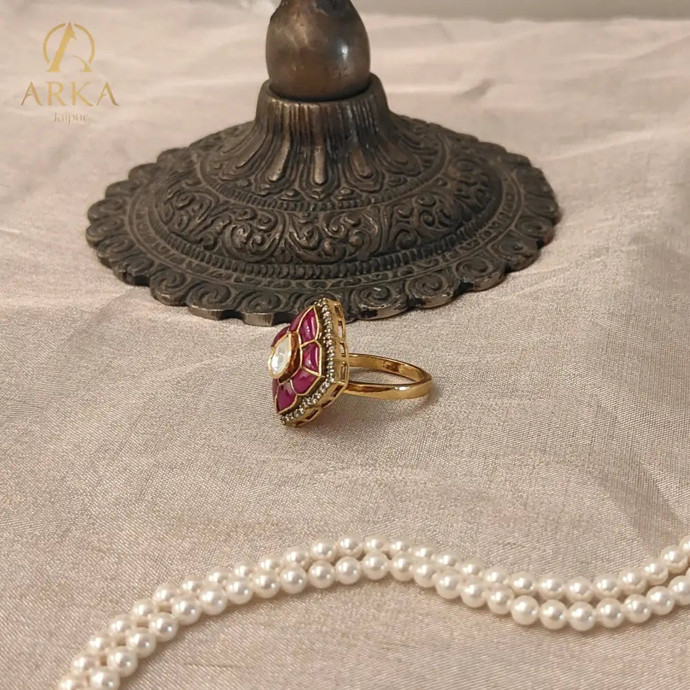 “Side view of the Lotus Bloom Ring by Arka Jaipur showing the raised lotus design in ruby enamel and gold finish, styled beside a brass decorative stand and pearl strand.”