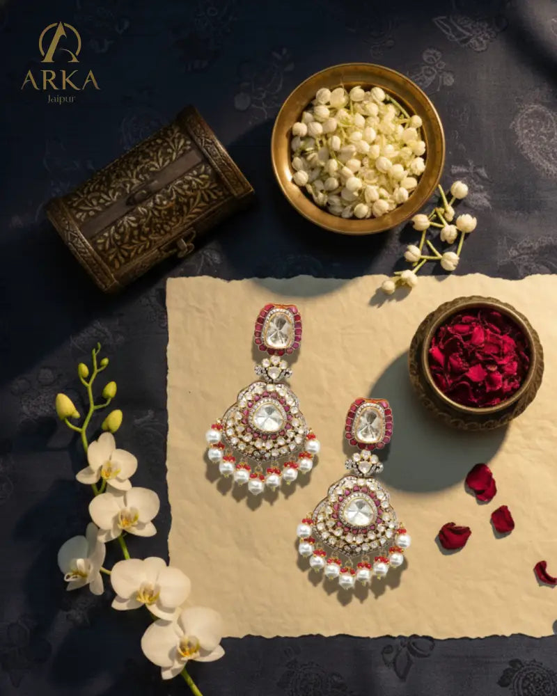 The Rosetta Royale earrings with pearls and red stones on a dark fabric background with flowers and a box.